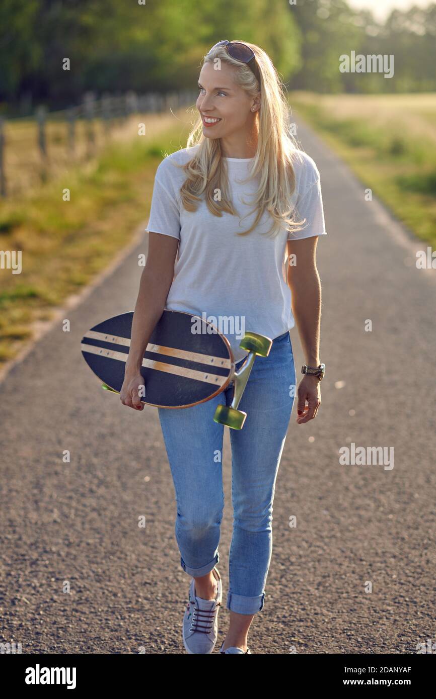 Fit gesunde junge blonde Frau trägt ein Skateboard entlang ein Schmale Landstraße Blick von der Seite mit einem glücklichen Lächeln von der Sonne beleuchtet Stockfoto