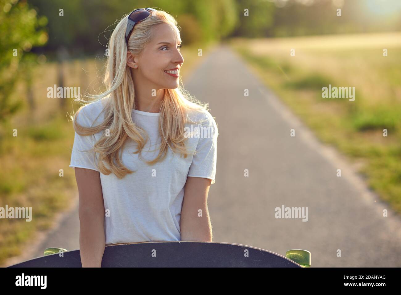 Fit gesunde junge blonde Frau trägt ein Skateboard entlang ein Schmale Landstraße Blick von der Seite mit einem glücklichen Lächeln von der Sonne beleuchtet Stockfoto