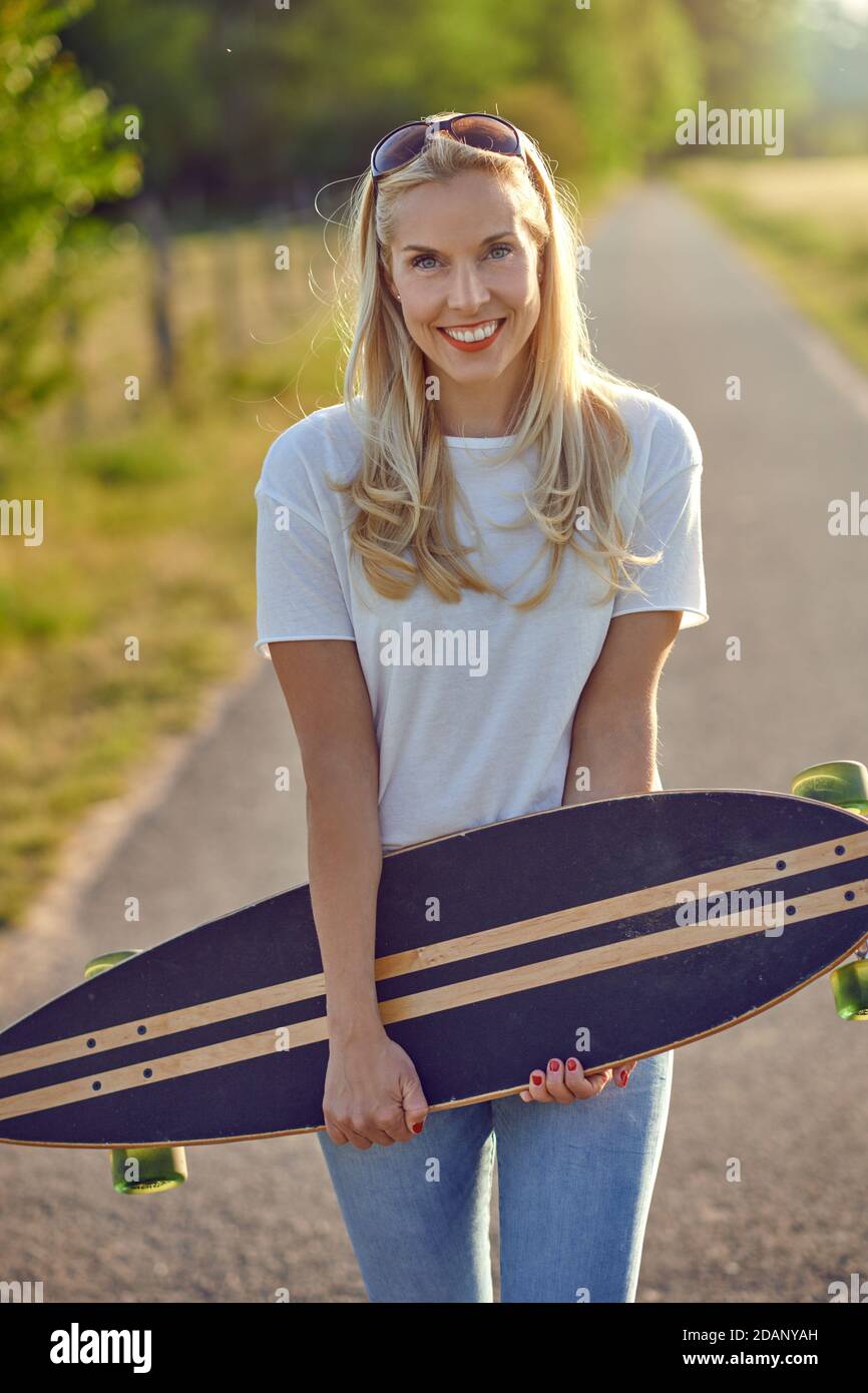 Porträt einer fit schöne Frau mittleren Alters mit einem aktiven Lebensstil lächeln und Blick auf die Kamera, während ein Longboard auf einer sonnigen Straße in der Stockfoto