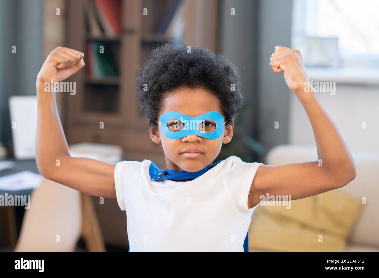 Niedliche Mixed-Rennen Junge in Kostüm von Superhelden zeigt Stärke in Vorderseite der Kamera Stockfoto