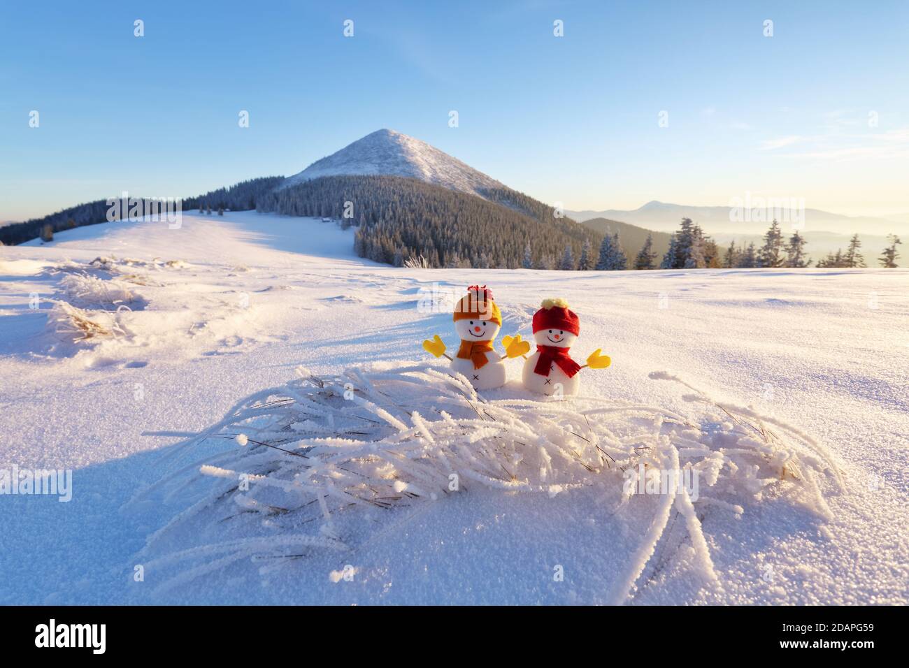 Paar zwei kleine Schneemänner in rotem Hut und Schal auf schneebedeckten Feld. Schöne Winter Sonnenuntergang Hintergrund. Landschaft mit hohen Bergen. Frohe weihnachten und Stockfoto