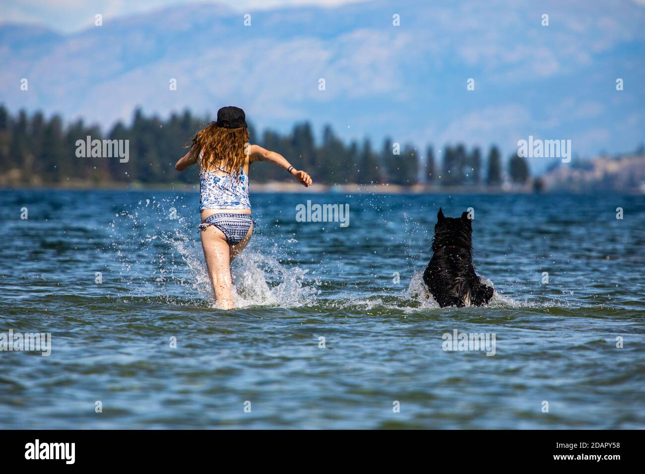 Langhaarige Frau und ihr Hund schossen von hinten, rannten vor der Kamera und spritzte knietief in das Wasser eines kanadischen Sees, aufsteigende Spritzer. Stockfoto