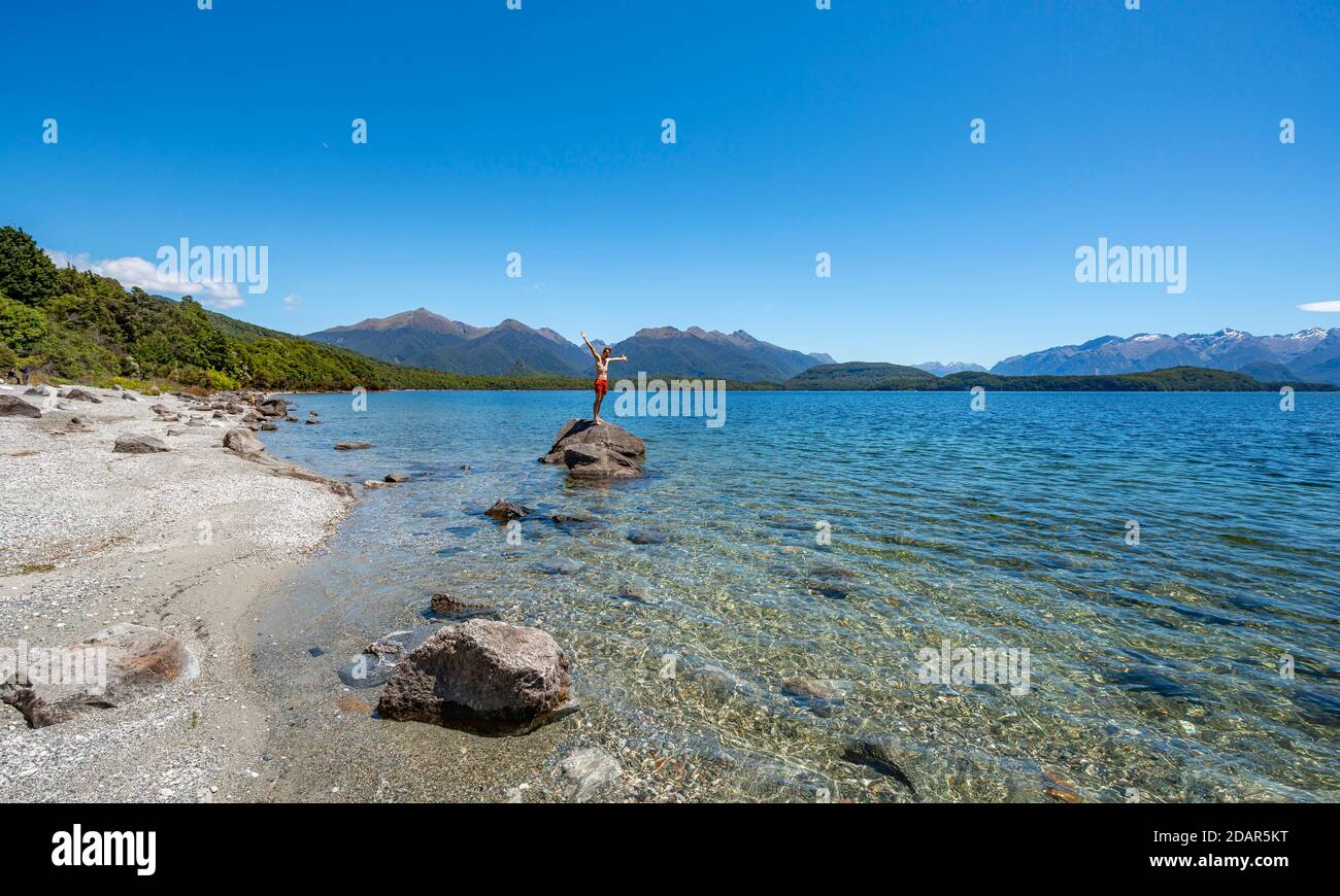 Junger Mann steht auf Stein im Wasser und streckt seine Arme in die Luft, Beach Fraser Beach, Lake Manapouri, Manapouri, South Island, Neuseeland Stockfoto