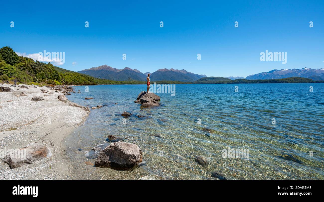 Junger Mann steht auf Stein im Wasser, Beach Fraser Beach, Lake Manapouri, Manapouri, South Island, Neuseeland Stockfoto