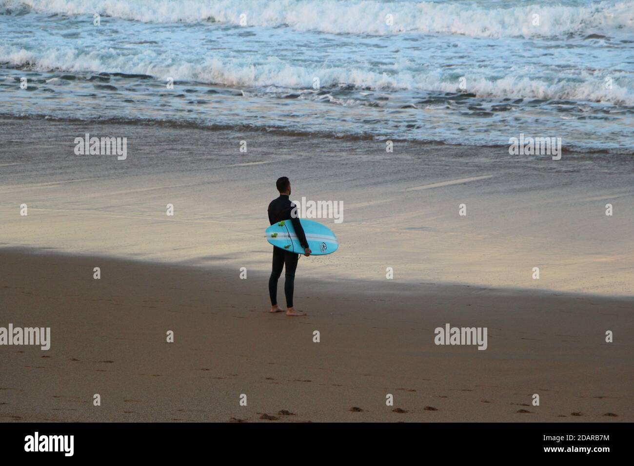 Surfer, der in Sardinero bei Sonnenuntergang am Strand mit seinem Surfbrett an einem milden Herbstabend steht, Santander Cantabria Spanien Stockfoto