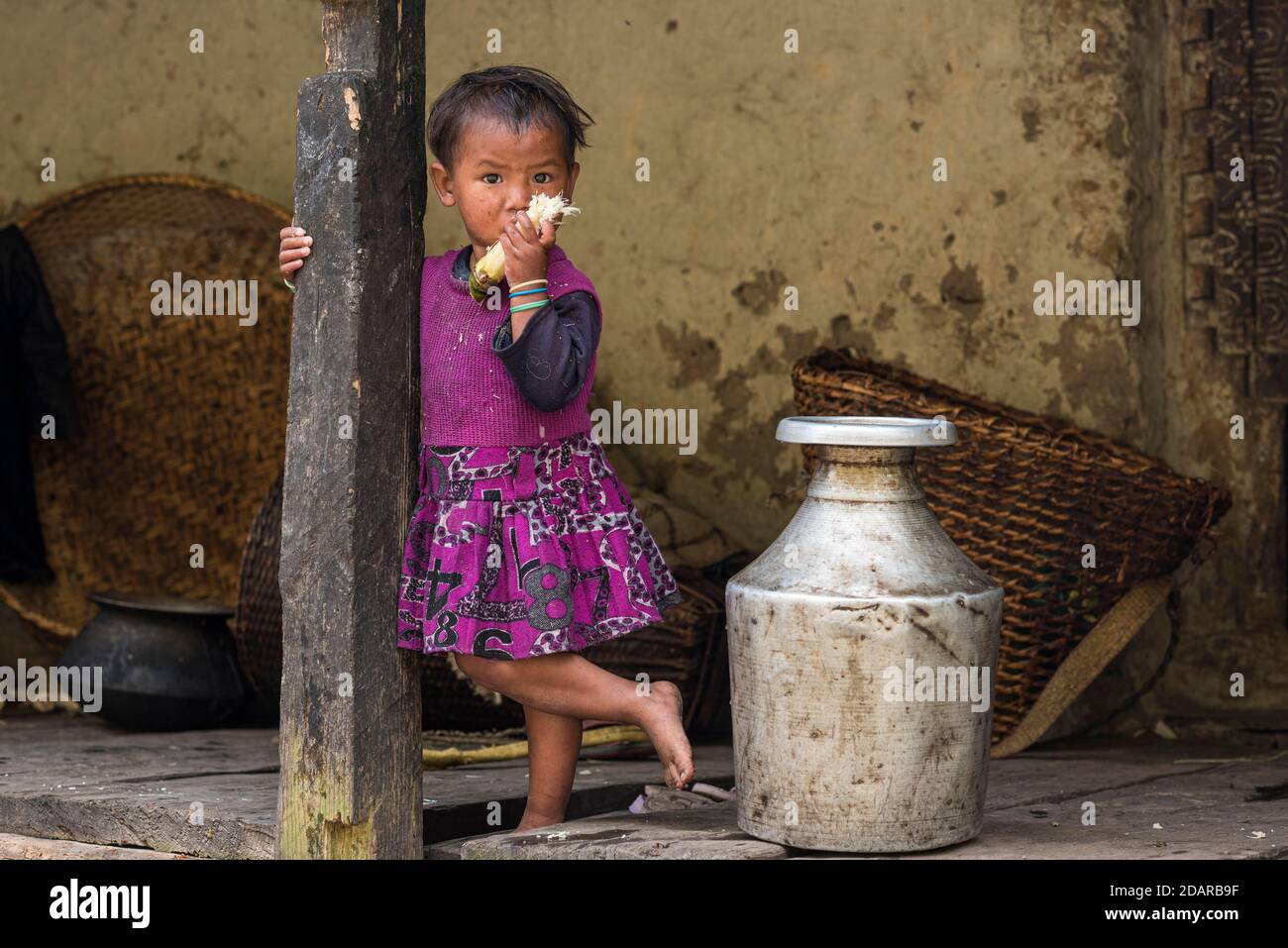 Ein kleines Mädchen steht vor dem Haus neben einem Aluminium-Wasserbehälter und kaut Zuckerrohr, Manaslu-Region, Himalaya, Nepal Stockfoto