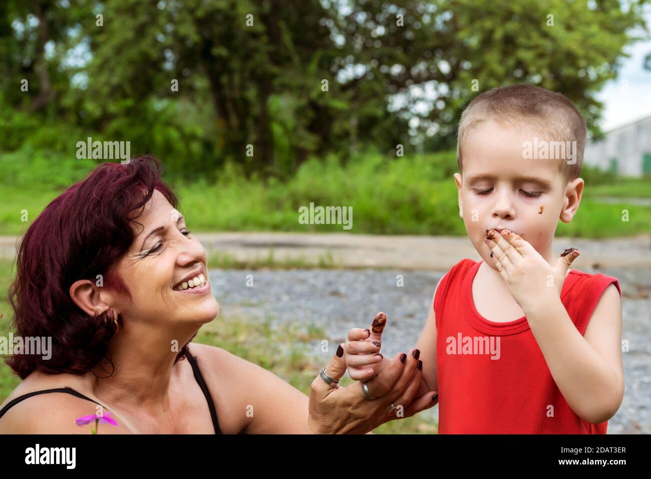 Ein Kind, das Schokolade direkt von seinen Fingern probiert, seine Großmutter ist direkt an seiner Seite und lächelt ihn an Stockfoto
