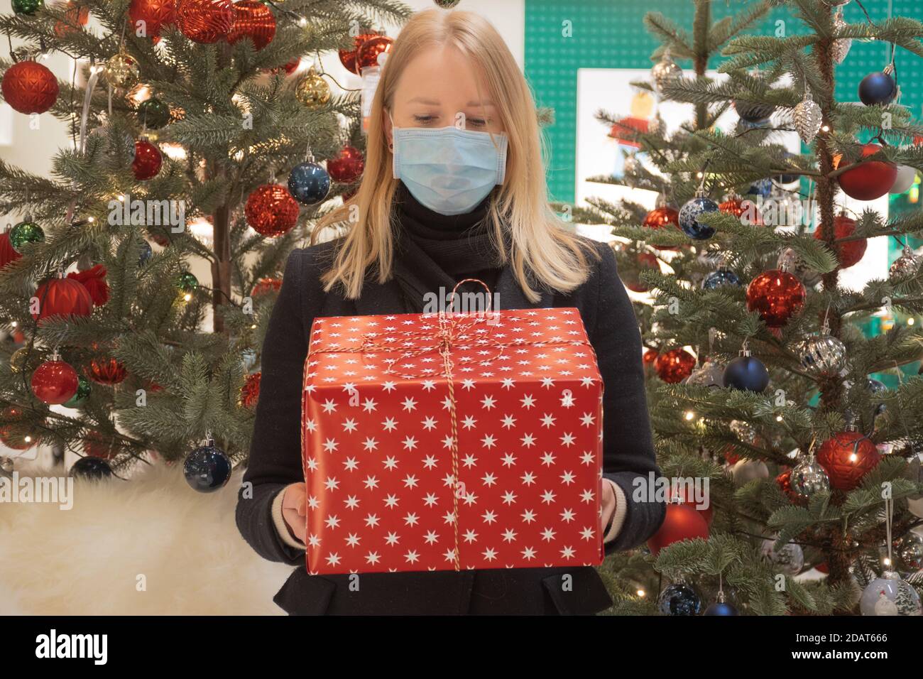 Schöne blonde Haare Mädchen mit schützenden Gesichtsmaske hält ein Geschenk oder Geschenk rote Box, Weihnachten Hintergrund mit Dekorationen und Bäumen Stockfoto