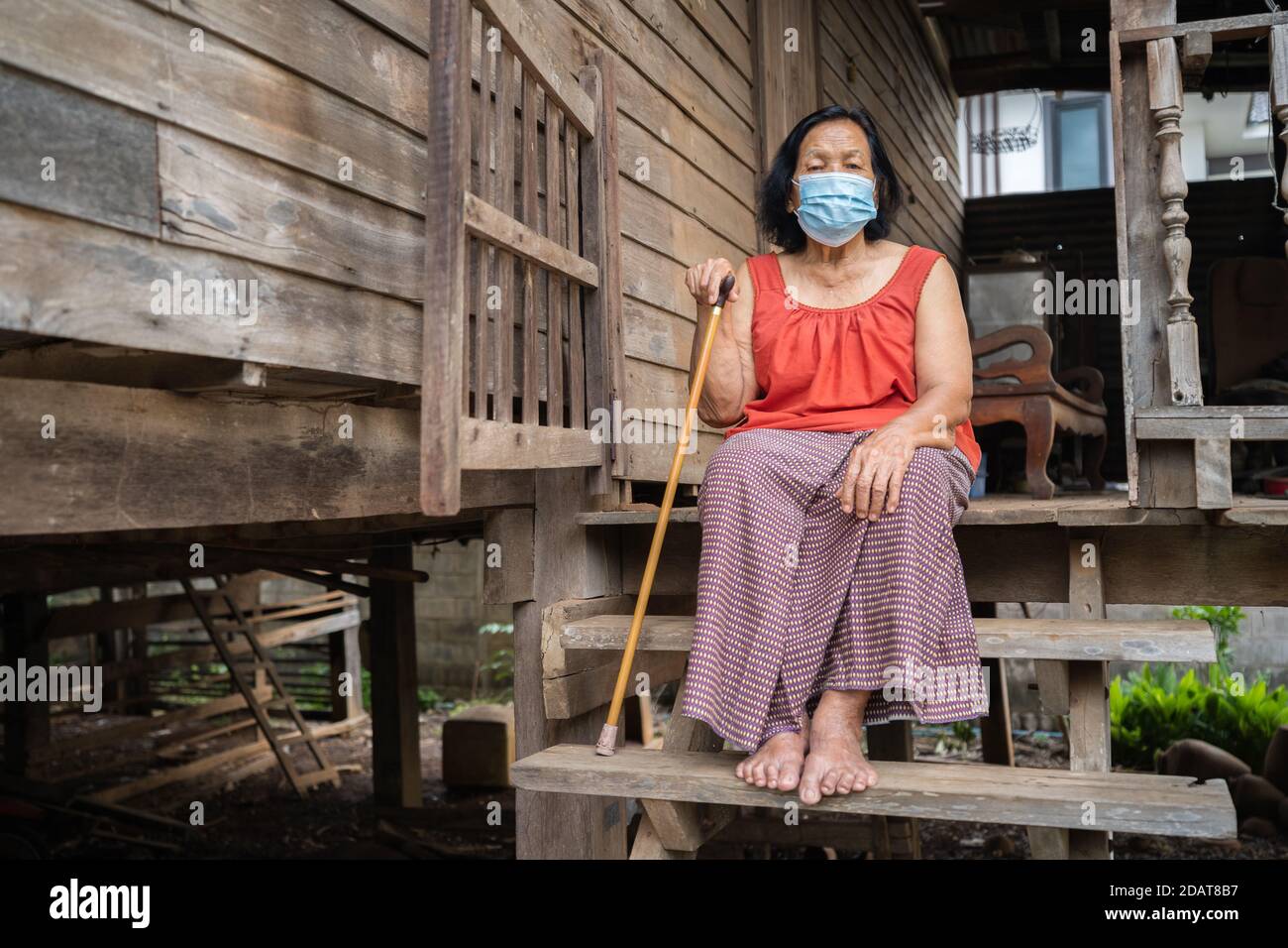 Thai ältere Frau in rund-Hals ärmellose Kragen trägt medizinische Maske Zum Schutz Corona Virus (covid-19) Pandemie in alten Holzhaus Stockfoto