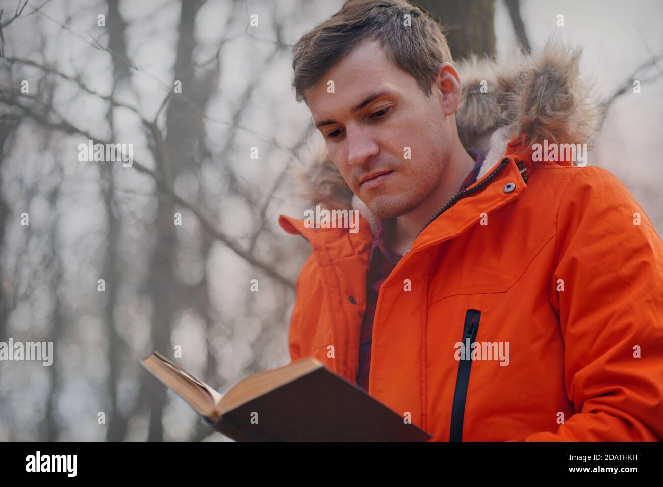 Erwachsene Mann in orange Jacke liest Buch im Herbst Wald. Junger Mann ruht mit Buch an der frischen Luft, genießt seine Freizeit Stockfoto