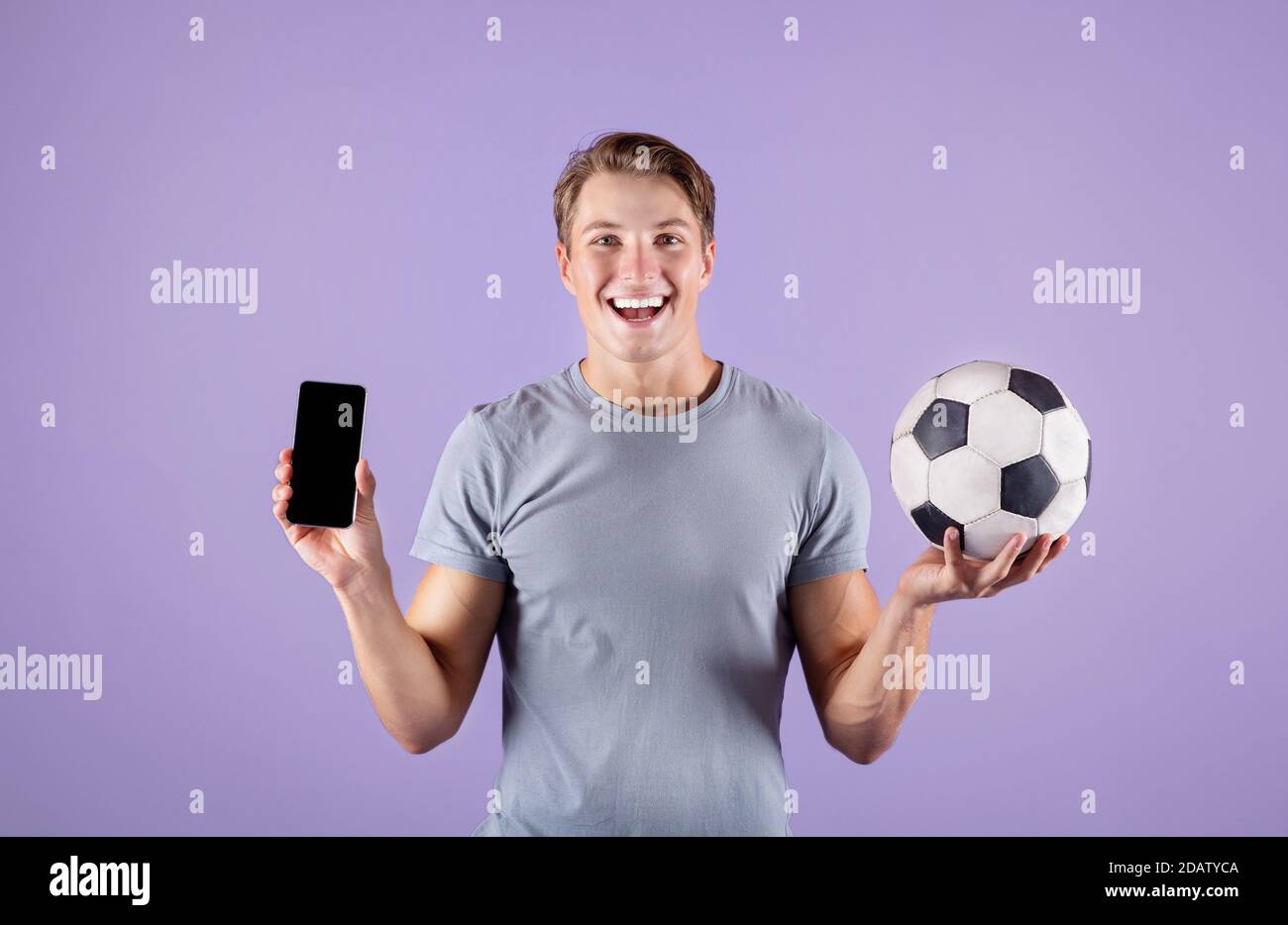 Happy Young guy hält Fußball und Smartphone mit leerem Bildschirm auf flieder Studio Hintergrund, mockup für Design Stockfoto