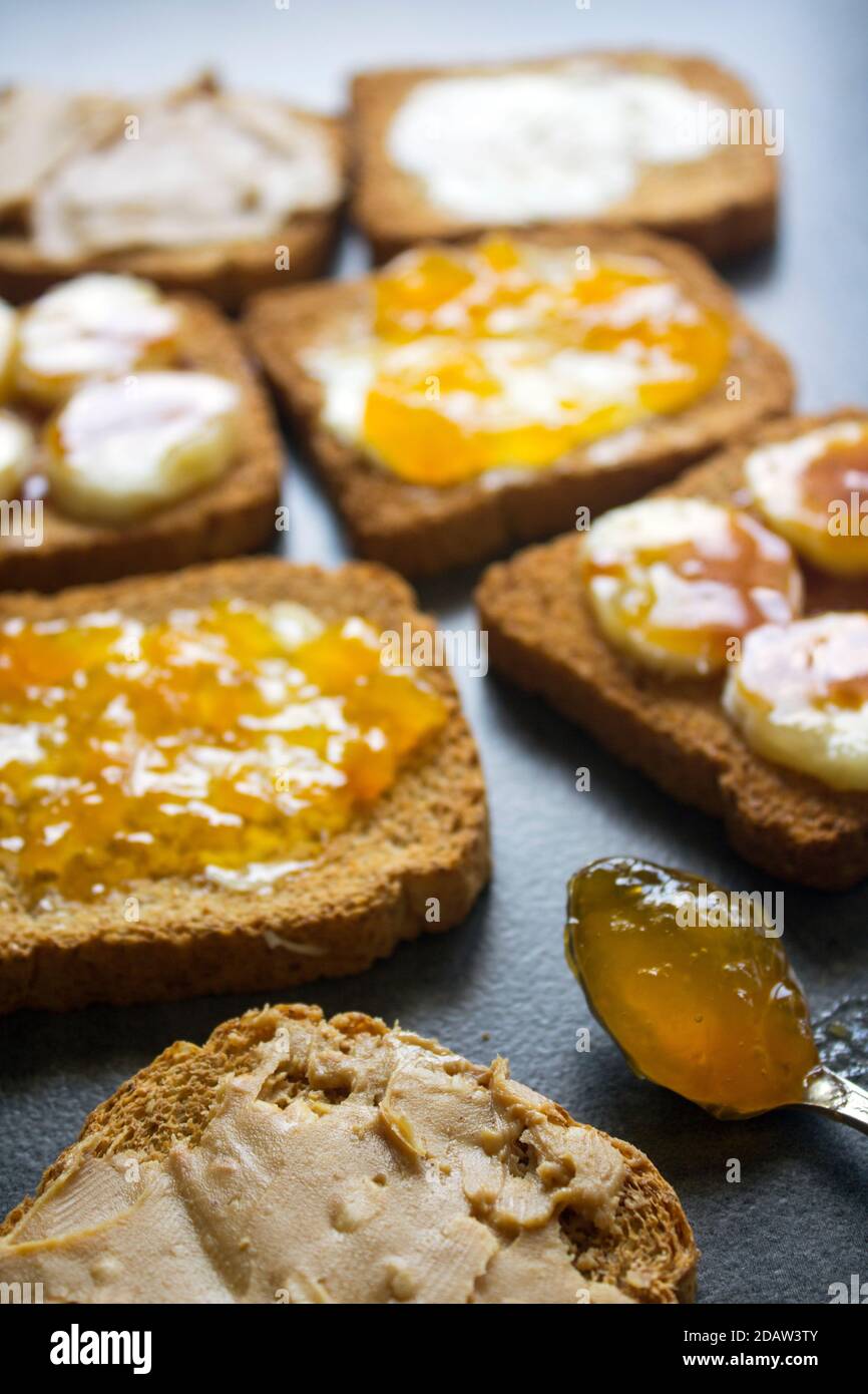 Süßes Frühstück Draufsicht Foto. Toasts mit verschiedenen Belägen: Erdnussbutter, Marmelade, Butter und karamellisierte Banane. Dunkelgrauer strukturierter Hintergrund Stockfoto