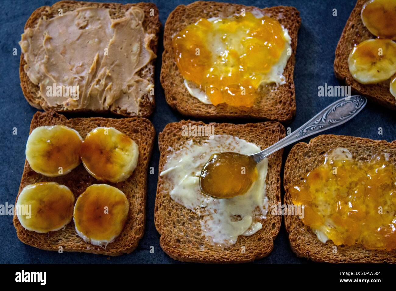 Süßes Frühstück Draufsicht Foto. Toasts mit verschiedenen Belägen: Erdnussbutter, Marmelade, Butter und karamellisierte Banane. Dunkelgrauer strukturierter Hintergrund mit Stockfoto