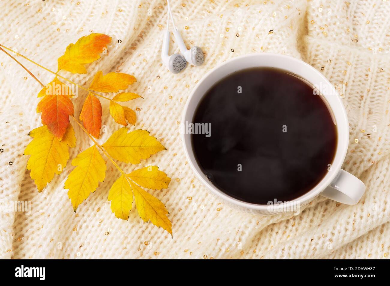 Großer weißer Becher mit heißem, dampfendem Kaffee, Ohrhörern und gelbem Herbstblatt auf einem beigefarbenen Strickschal. Gemütliches Herbststimmung Konzept. Draufsicht. Stockfoto