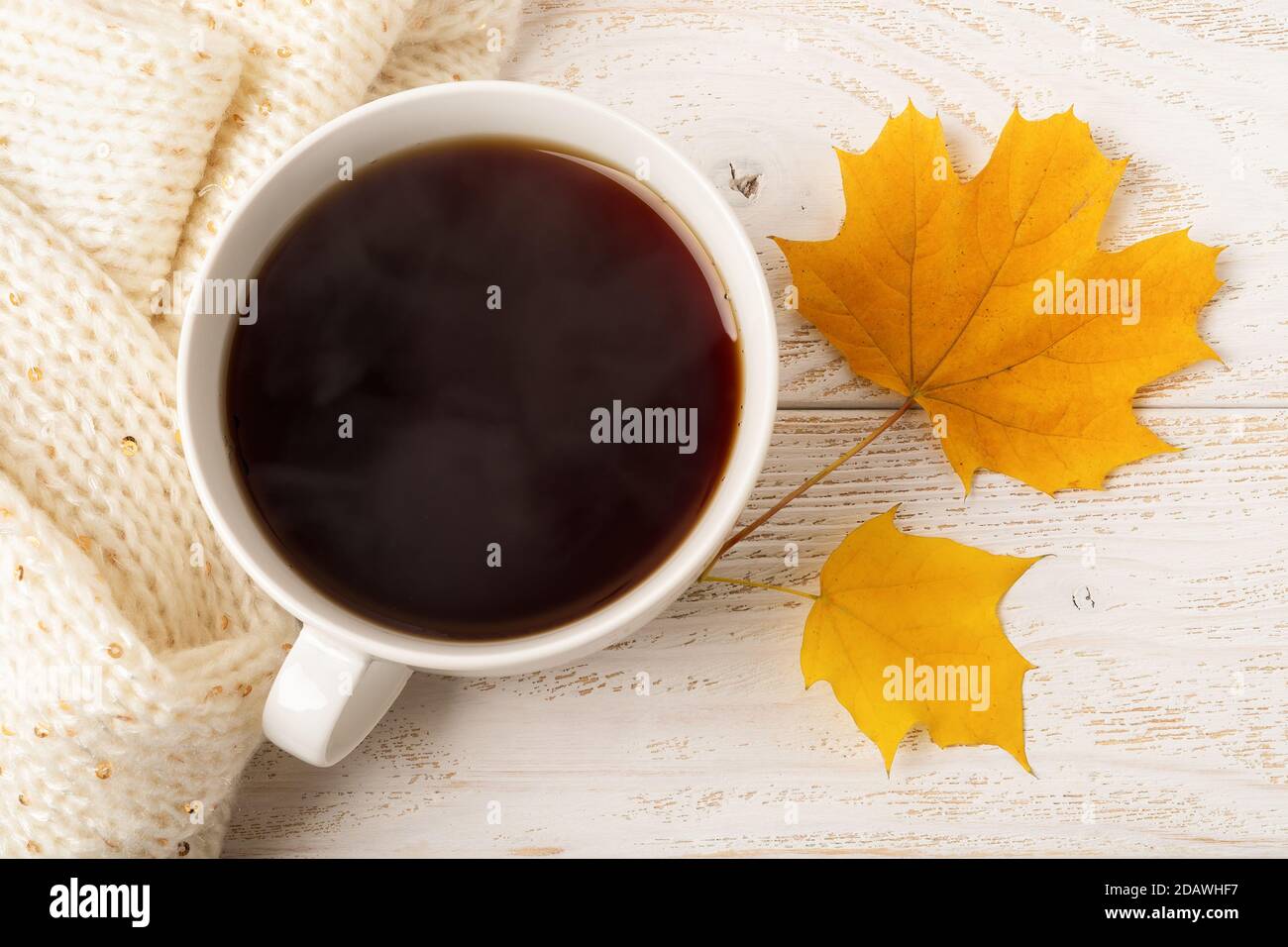 Große weiße Tasse heißen dampfenden Kaffee und zwei gelbe Herbstblätter in der Nähe beige gestrickten Schal über weißem Holz rustikalen Oberfläche. Gemütliches Herbstkonzept. Stockfoto