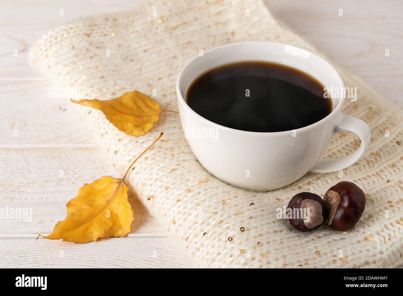 Große weiße Tasse heißen dampfenden Kaffee auf einem beigen Strickschal und zwei gelben Herbstblätter über weißem Holz rustikale Oberfläche. Gemütliches Herbststimmung Konzept. Stockfoto
