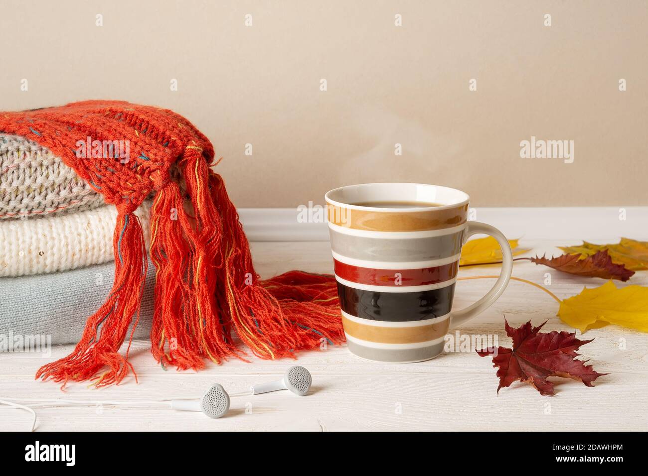 Gestreifte Tasse heißen dampfenden Kaffee oder Tee, Stapel von warmen Strickpullover und rot Fransen Schal und einige gelbe Herbstblätter über Holz rustikalen Oberfläche. Stockfoto