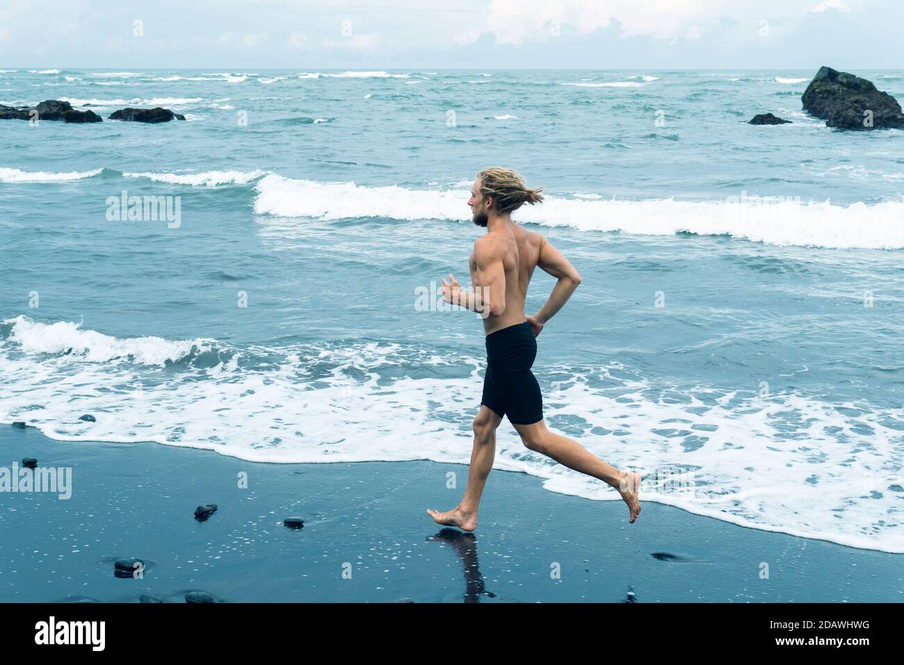 Sportler Mann Joggen auf einem schwarzen Sandstrand mit Meer Hintergrund Stockfoto