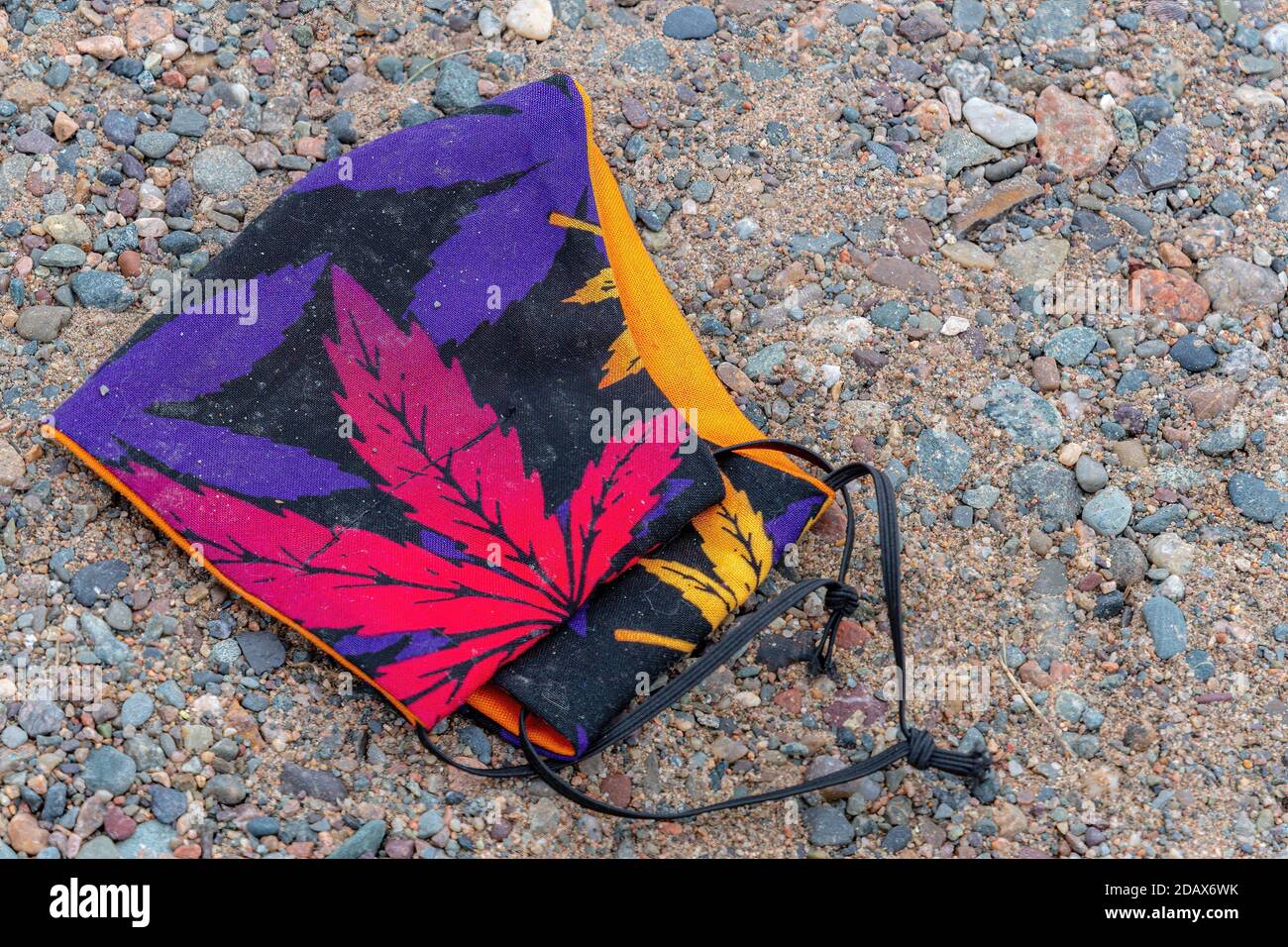 Eine Maske mit roten und violetten Cannabisblättern. Maske liegt auf Kies. Dies ist die Art der Maske, die Menschen verwenden, um die Ausbreitung des Coronavirus zu verhindern. Stockfoto