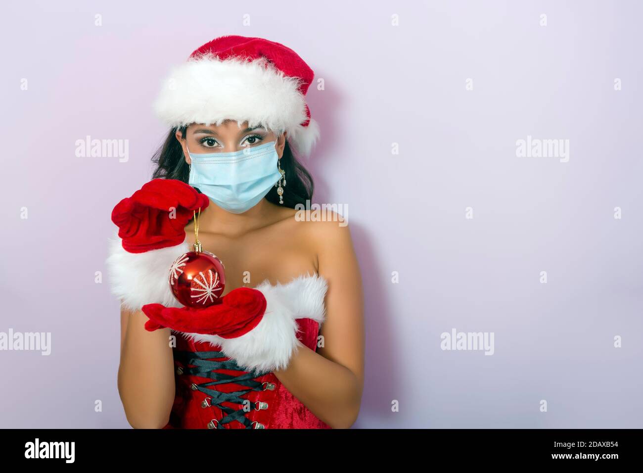 Junge Brünette Frau in santa claus Kostüm und chirurgische gekleidet Maske mit weihnachtskugel in den Händen Stockfoto