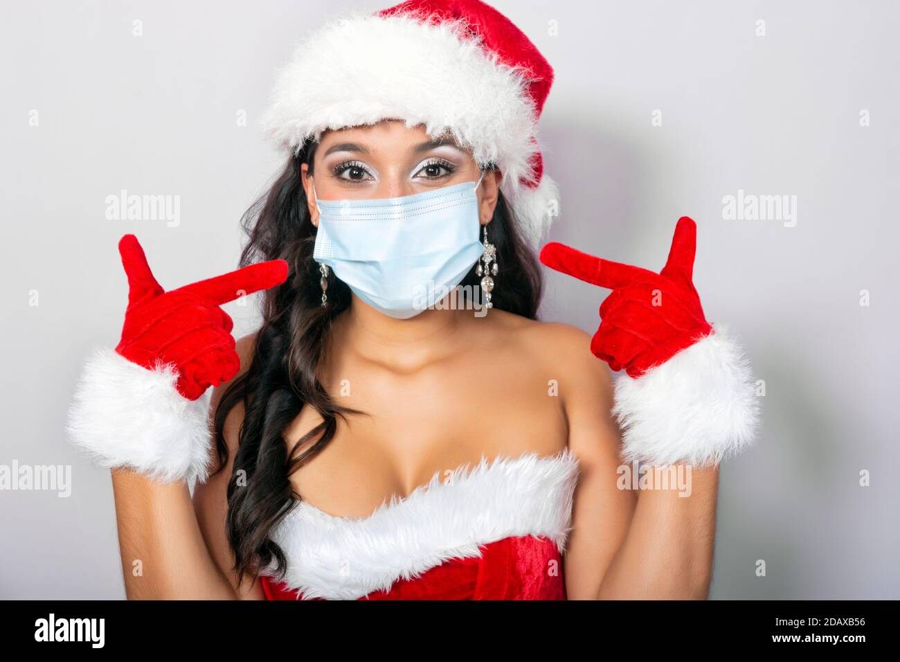 Junge Brünette Frau in weihnachtsmann Kostüm gekleidet zeigt auf Ihre Anti-Virus-Schutzmaske Stockfoto