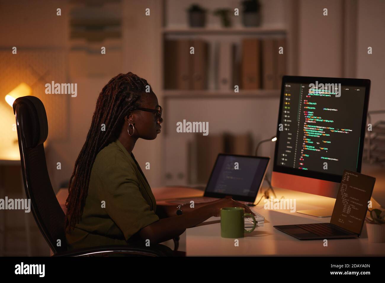Seitenansicht Porträt einer zeitgenössischen afroamerikanischen Frau, die Code schreibt und auf den Computerbildschirm schaut, während sie in einem dunklen Büro arbeitet, Kopierraum Stockfoto