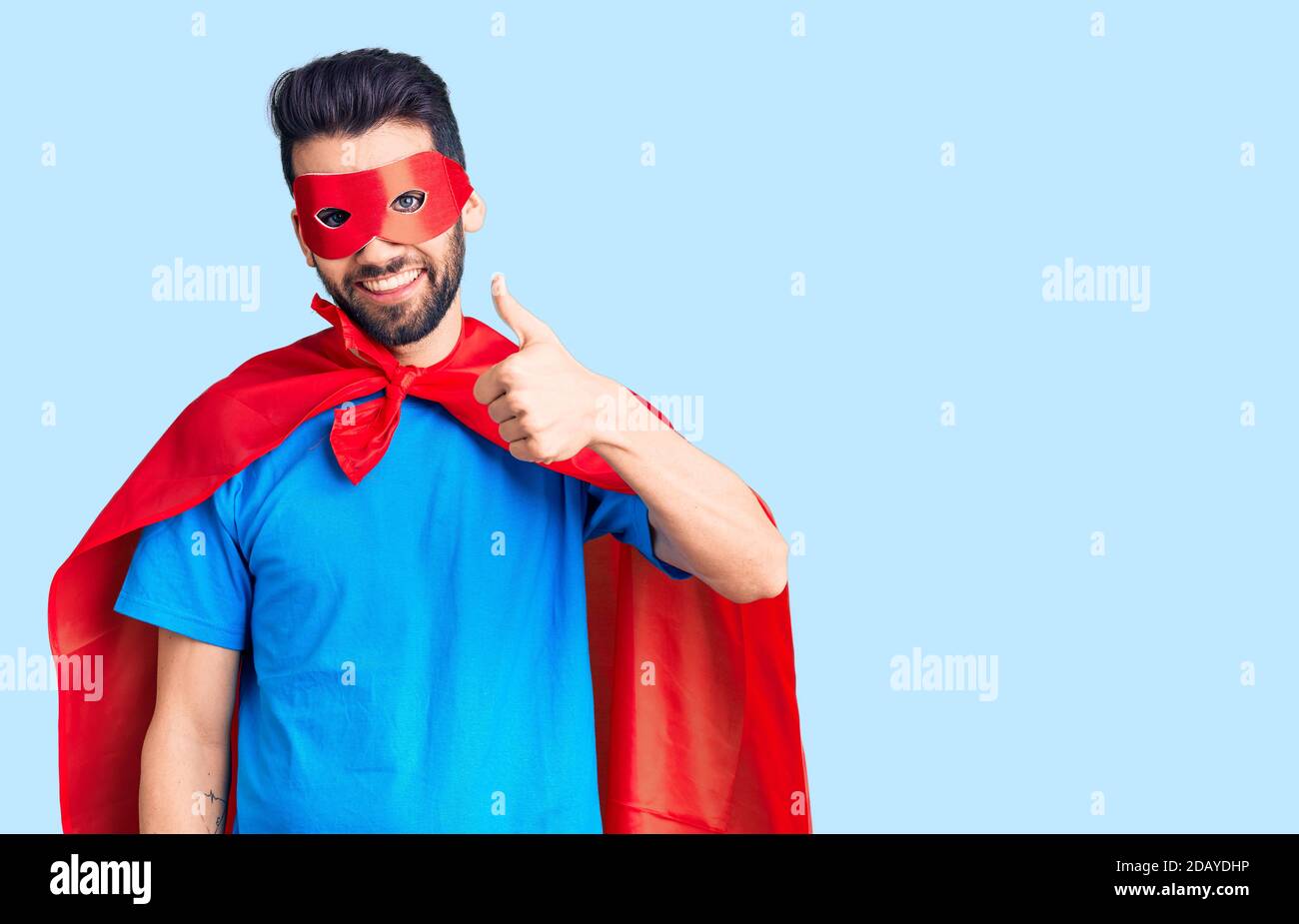 Junger schöner Mann mit Bart trägt Superhelden Kostüm tun glückliche Daumen nach oben Geste mit der Hand. Genehmigung Ausdruck Blick auf die Kamera zeigt s Stockfoto
