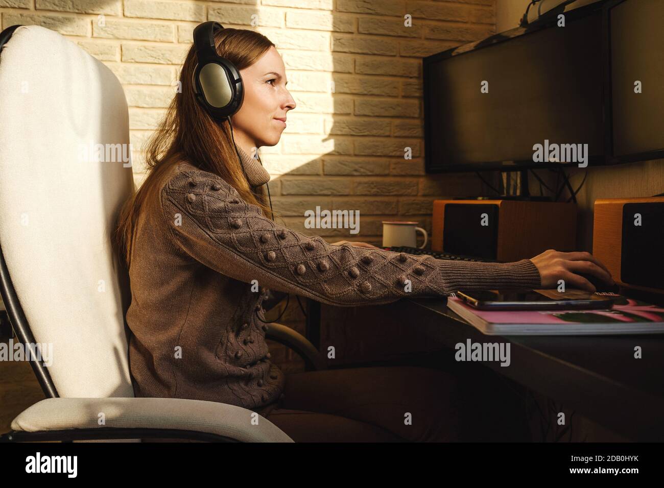 Junge Frau in Kopfhörer mit Videoanruf über Computer zu Hause. Remote-Arbeit, Sonnenlicht an der Wand. Leerer Bildschirm für Ihr Foto. Stockfoto