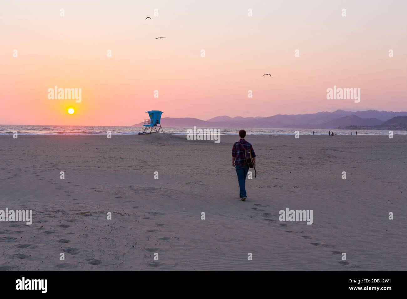 Junger Mann mit Gitarre zu Fuß in Richtung blauen Rettungsschwimmer Turm an Sunset Pismo Beach Kalifornien Stockfoto
