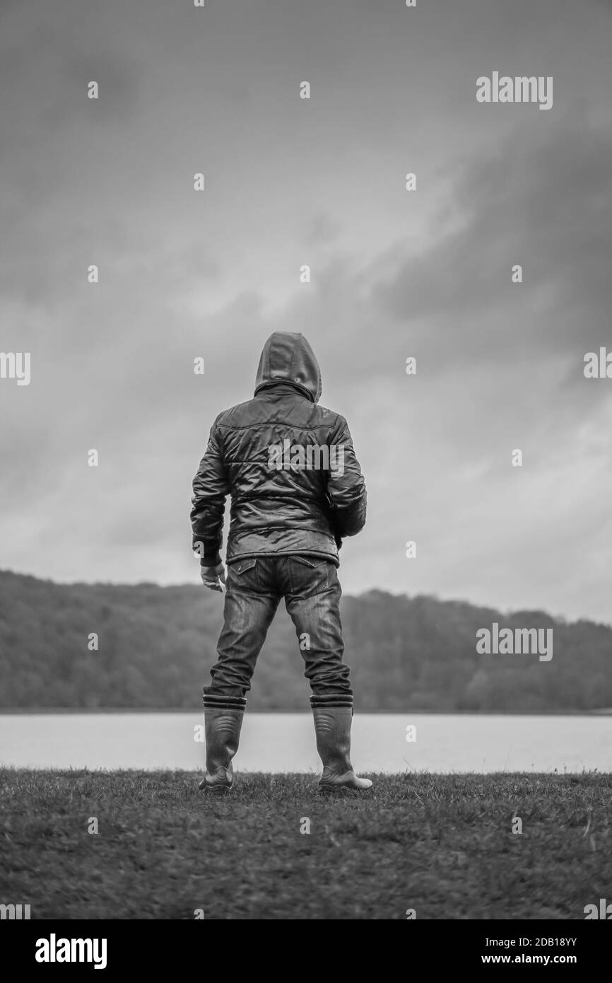 Porträt, mono, Rückansicht eines isolierten Mannes, der in Gummistiefel steht, Mantel mit Kapuze und Blick über Wasser mit launischen Wolken darüber; bedrohliches Aussehen. Stockfoto