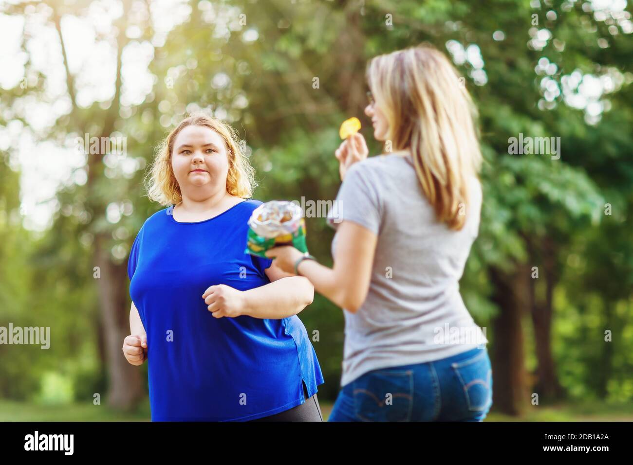 Eine junge Blondine von plus Größen läuft im Park in der Nähe einer Frau, die Chips isst. Das Konzept eines gesunden Lebensstils und der Selbstverbesserung Stockfoto