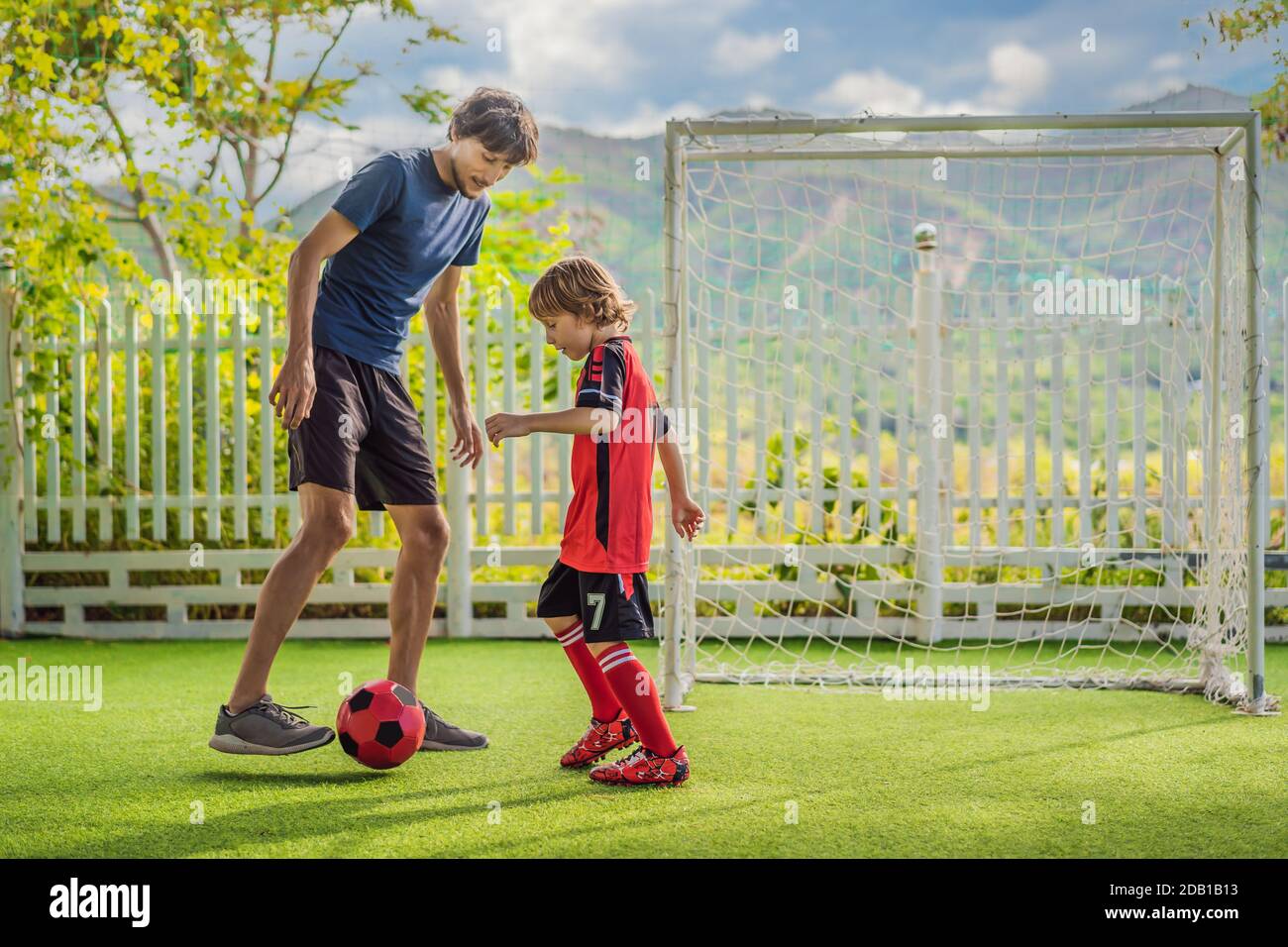 Kleiner niedlicher Junge in roter Fußballuniform und sein Trainer oder Vater spielt Fußball, Fußball auf dem Feld, im Freien. Aktives Kind macht Sport mit Stockfoto