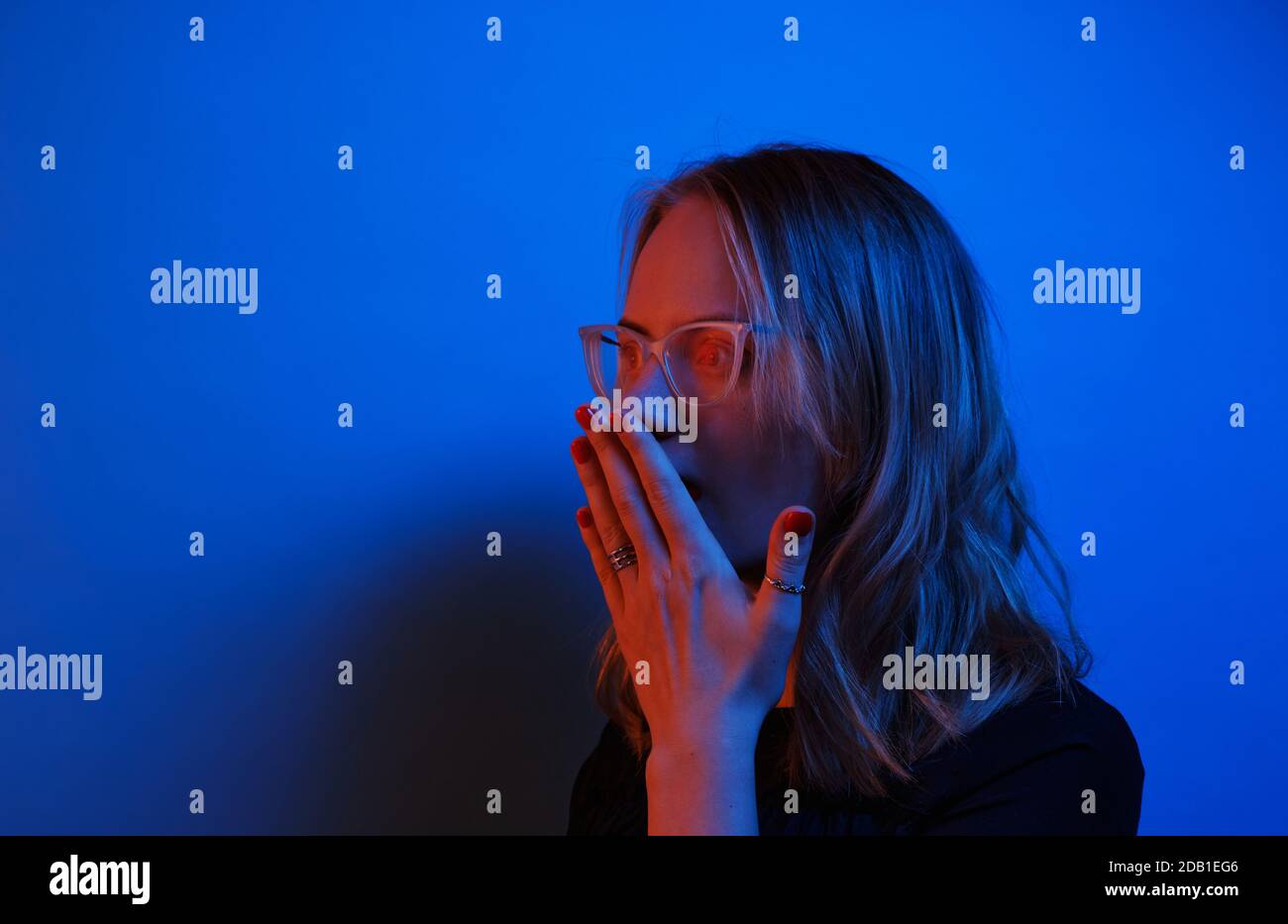 Ein junges kaukasisches Mädchen in Brille sieht auf einem dunkelblauen Neon-Hintergrund überraschend zur Seite. Menschliche Emotion Konzept und Raum kopieren. Stockfoto