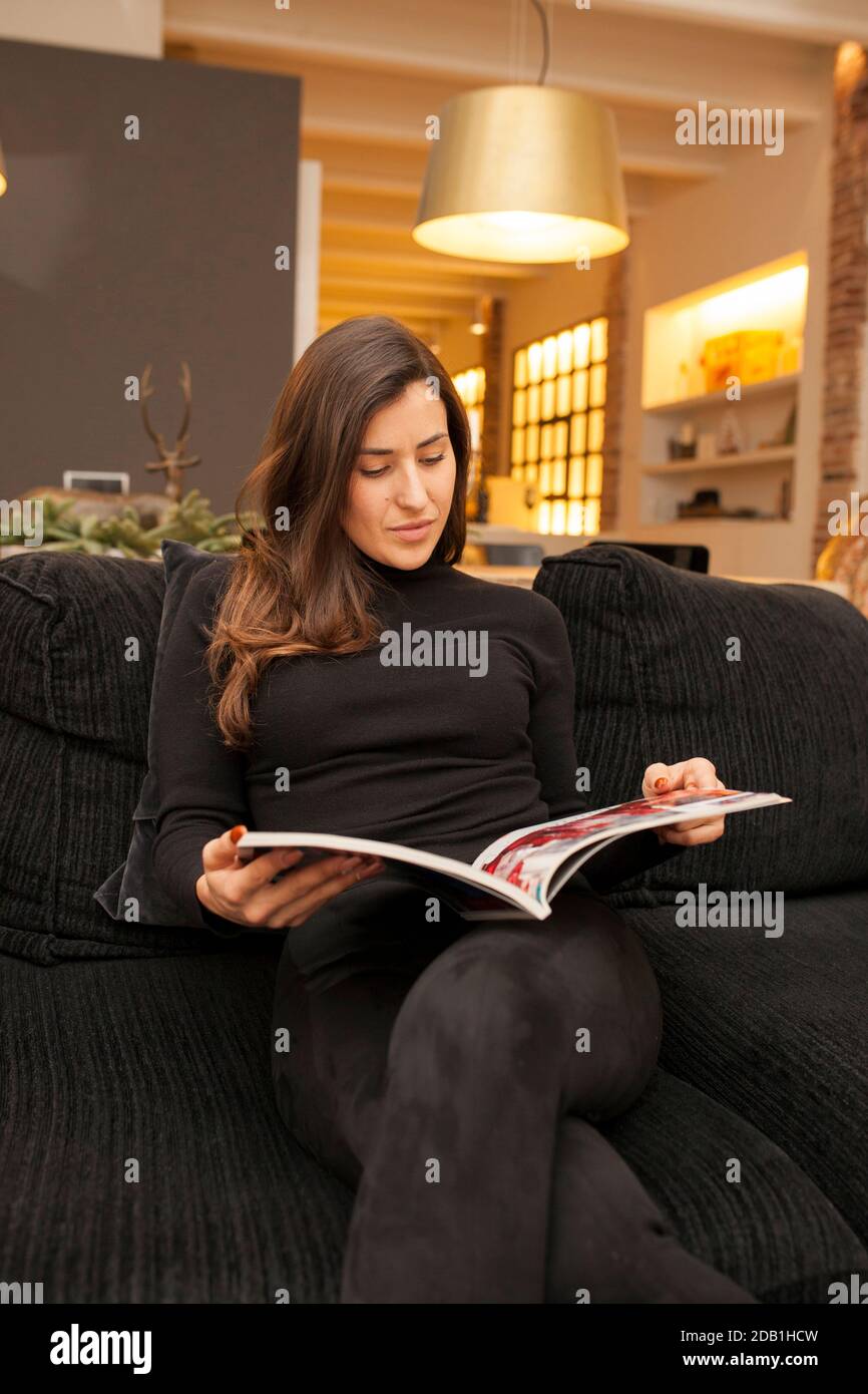 Eine schöne kaukasische Frau, die einen schwarzen Rollkragen trägt und einen liest Magazin auf dem Sofa Stockfoto