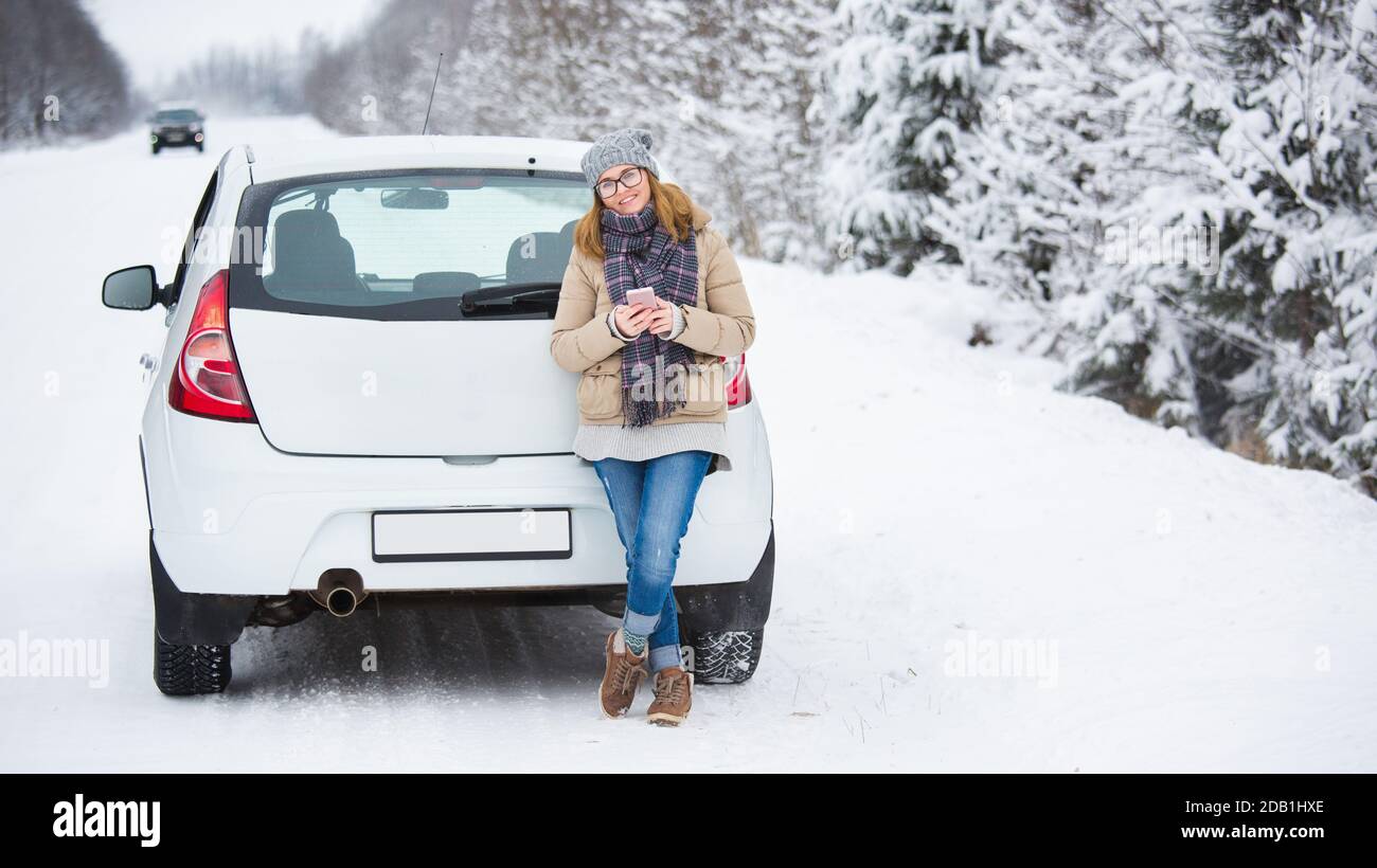 Frau mit Smartphone steht neben dem Auto auf einem Hintergrund Des schneebedeckten Winterwaldes Stockfoto