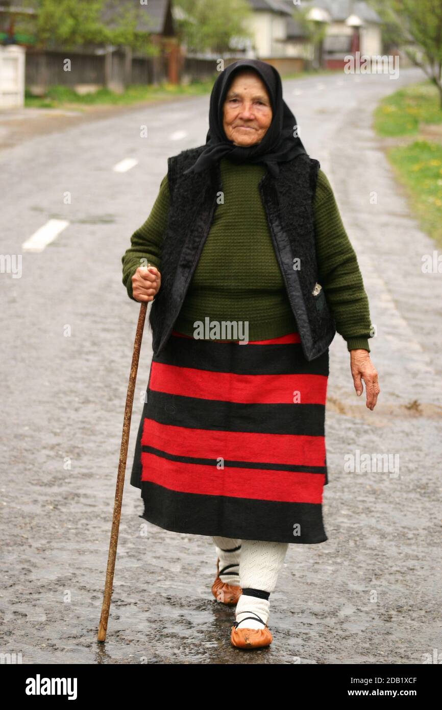Maramures, Rumänien. Ältere Frau in traditioneller Kleidung, die mit einem Stock auf der Dorfstraße läuft. Stockfoto