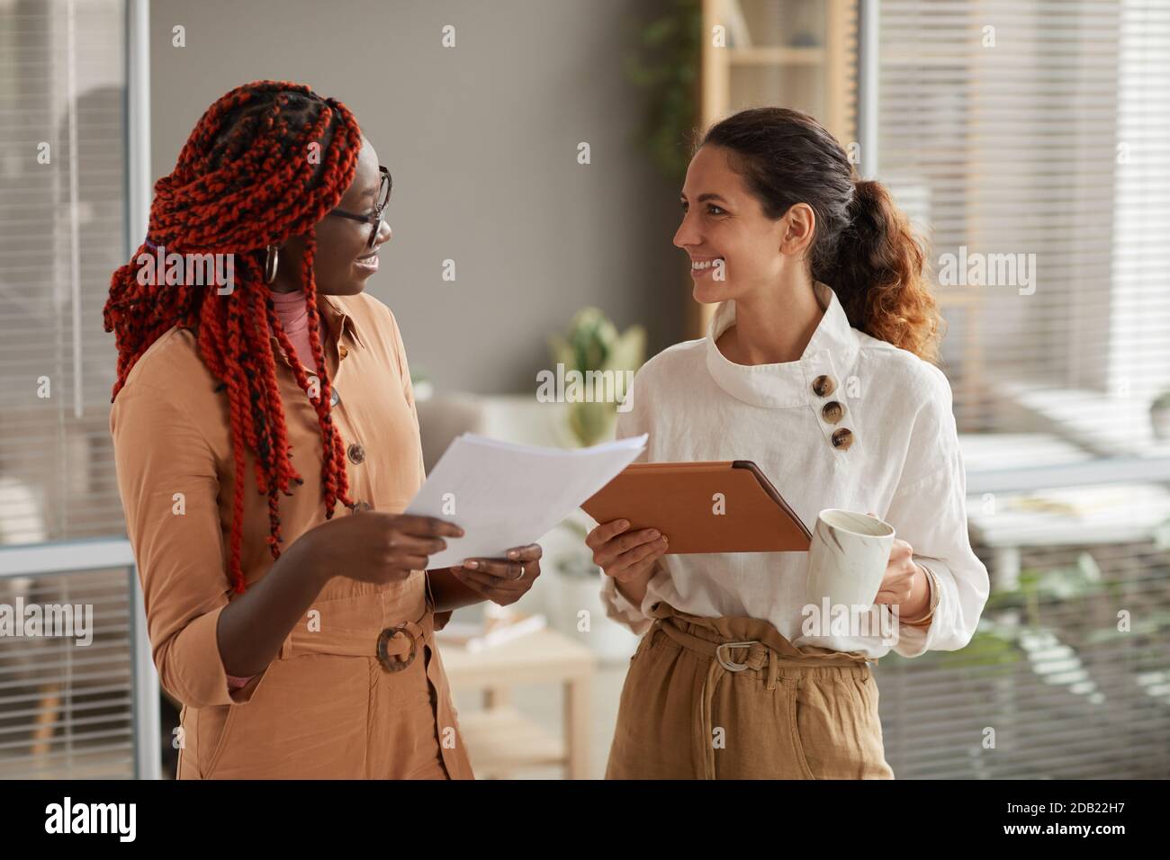 Waist-up-Porträt von zwei erfolgreichen jungen Geschäftsfrauen diskutieren Dokumente und lächeln fröhlich, während im modernen Büroinnenraum, Kopierer Raum stehen Stockfoto