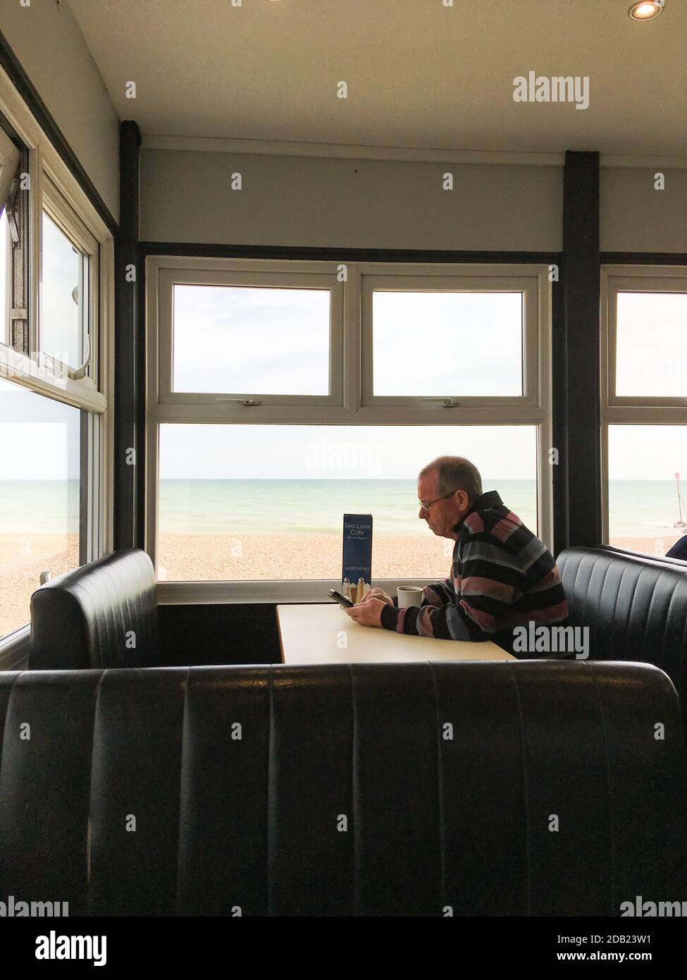 Ein Mann mittleren Alters sitzt allein in einem Café mit einer Tasse Kaffee SMS auf seinem Handy mit Blick auf das Meer in West Sussex, England, Großbritannien. Stockfoto