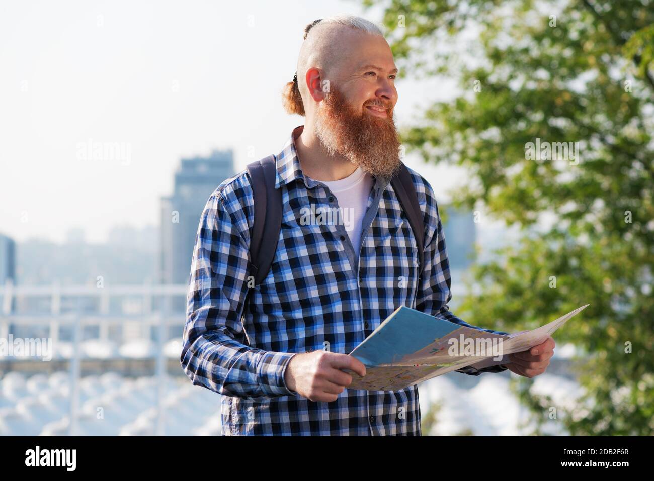 Lächelnder kaukasischer Hipster bärtiger Mann, der mit einem aktiven Urlaub zufrieden war. Lächelnder männlicher Reisender, der am Wochenende mit einer Karte durch die Stadt spazierenging Stockfoto