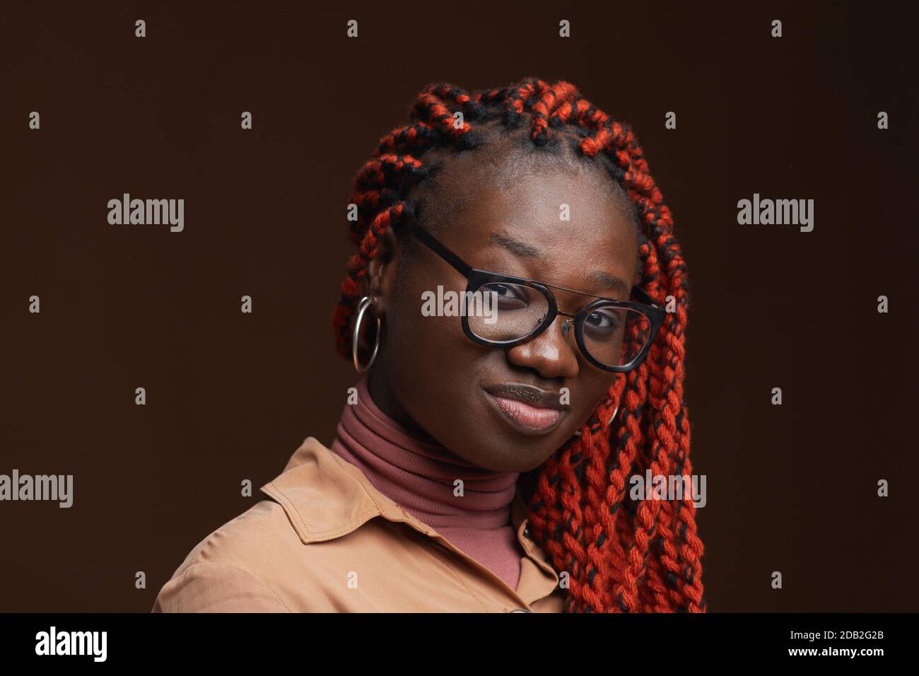 Nahaufnahme Porträt der zeitgenössischen afroamerikanischen Frau mit geflochtenen Haaren Blick auf die Kamera, während posiert vor dunkelbraunem Hintergrund im Studio, Copy Space Stockfoto