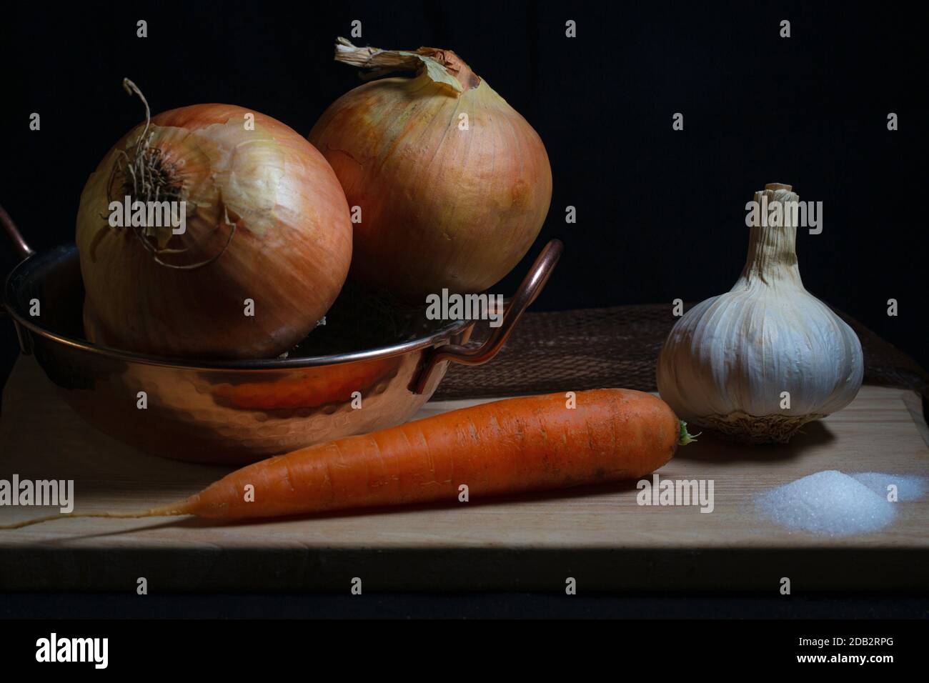 Stillleben Bild von Zwiebeln, Karotten und Knoblauch mit einer Prise Salz über Holz Schneidebrett mit einem schwarzen Hintergrund Stockfoto