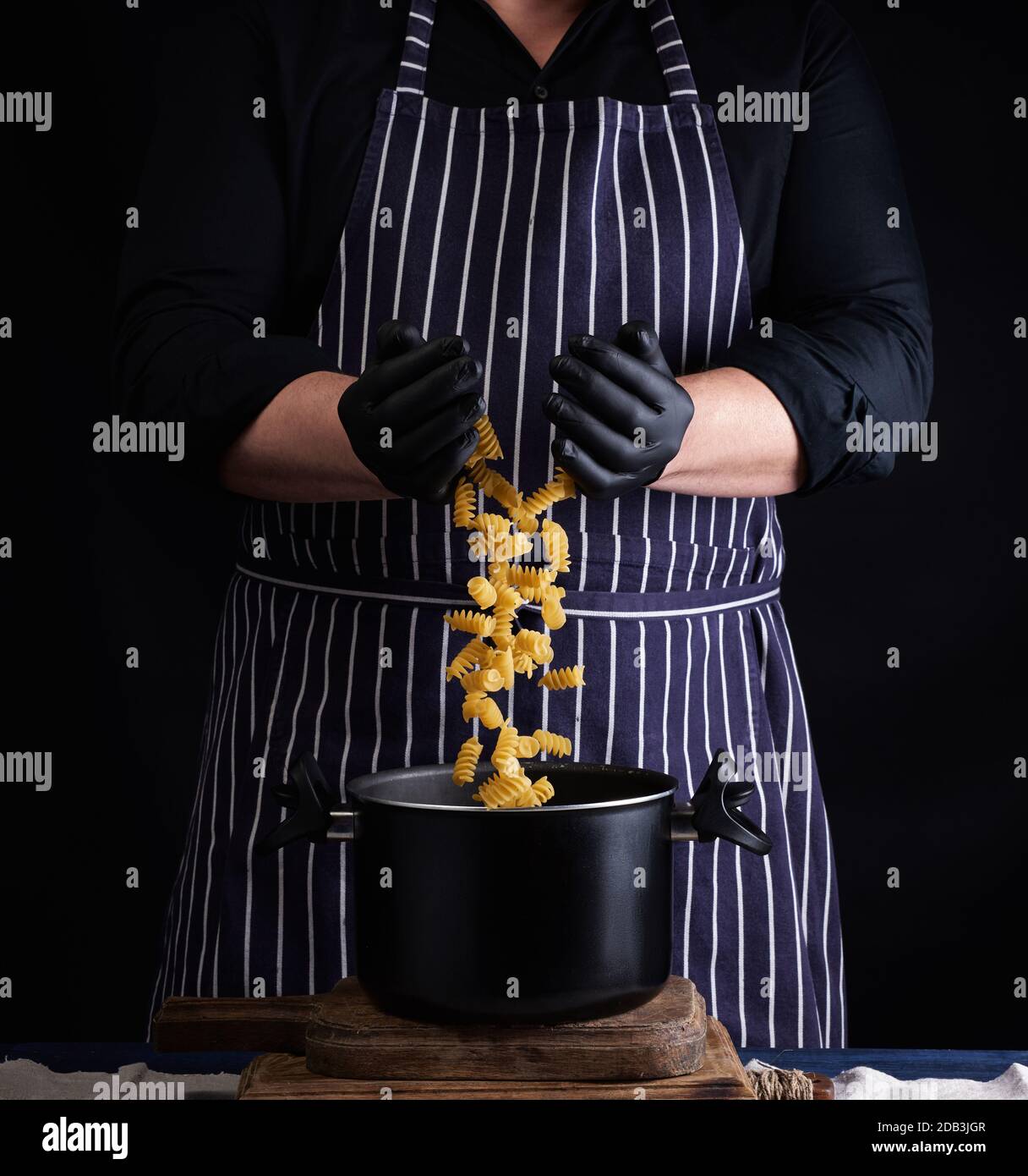 Chefkoch in schwarzen Latexhandschuhen, gestreifte Schürze gießt rohe Fusilli-Pasta in eine Metallpfanne, Kochvorgang, verdrehte Pasta in der Luft gefroren Stockfoto