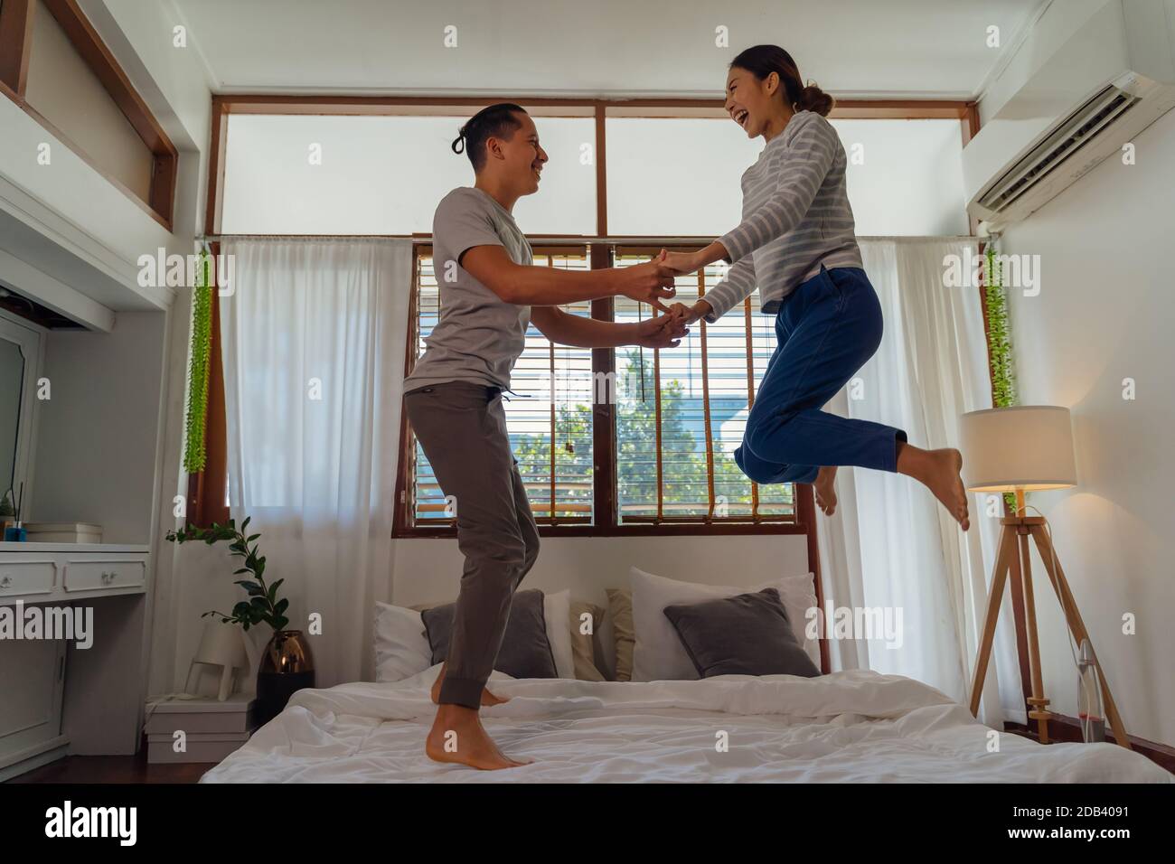 Portrait von glücklich junge Erwachsene asiatische Paar springen auf Bett zusammen im Schlafzimmer Innenleben Szene. 30s reifer Ehemann und Ehefrau lächeln und halten Hände. Ehe und glückliche Beziehung Leben Konzept Stockfoto