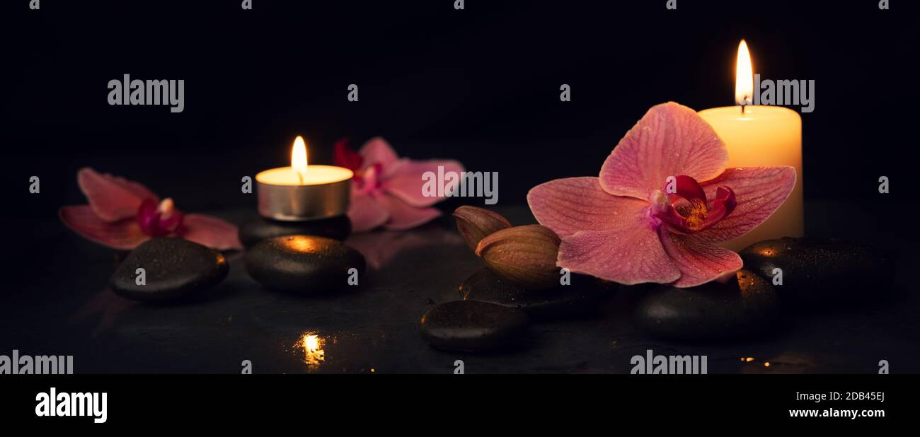 Spa und alternative Therapie. Kerzen mit Massagesteinen und Orchideenblumen auf schwarzem Hintergrund. Banner Stockfoto