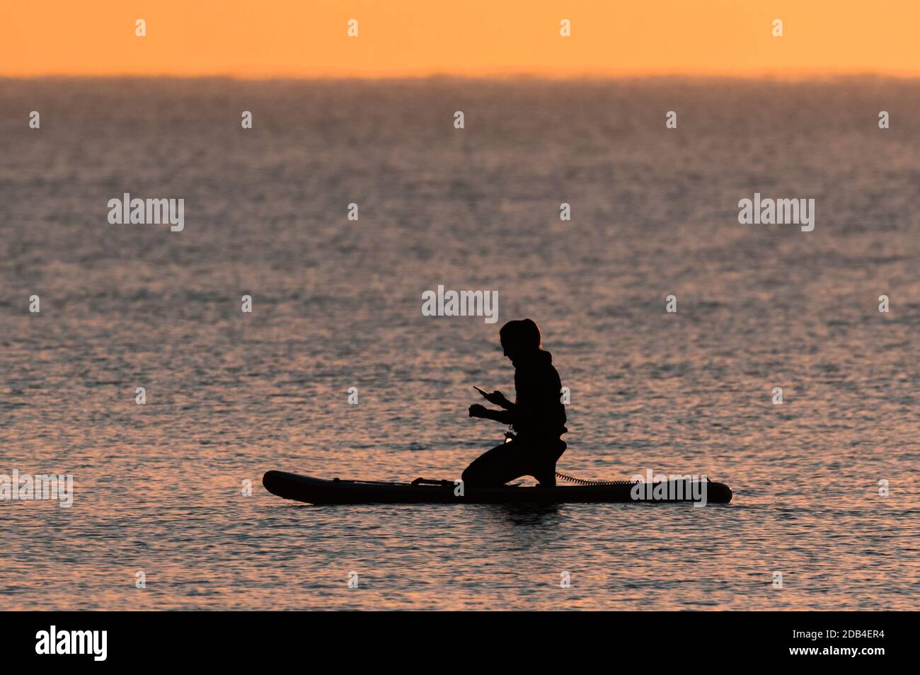 Silhouette einer Frau auf einem Paddleboard auf dem Meer in wenig Abendlicht, mit einem Handy. Stockfoto
