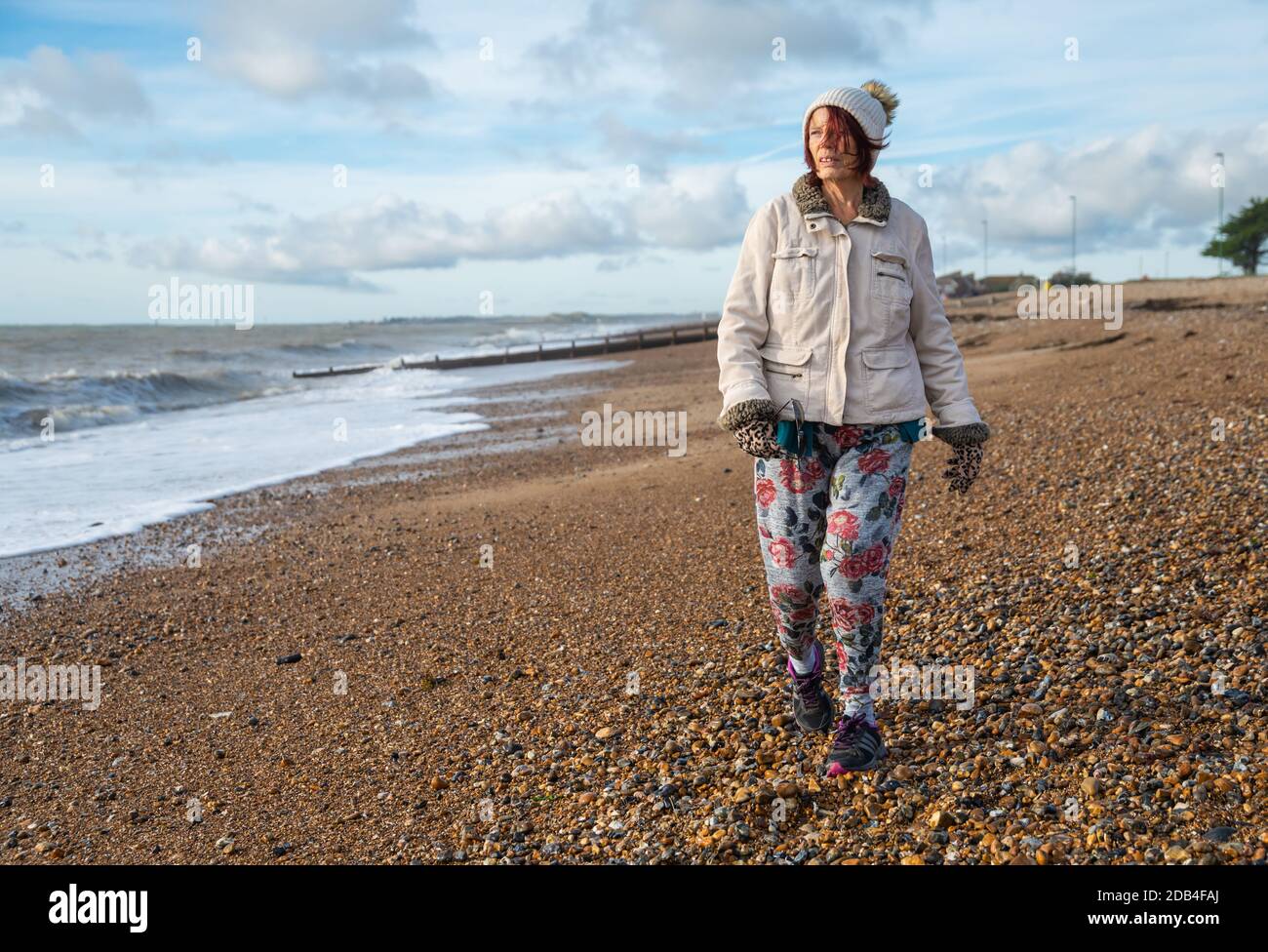 Ältere Frau in Mantel, Hut, Handschuhe und Hose, zu Fuß auf einem Strand am Meer im Herbst in Großbritannien. Strandpromenade. Stockfoto