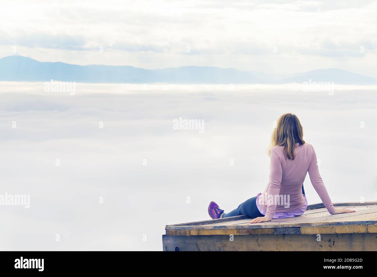 Frau in sportlicher Kleidung, die auf einer hölzernen Plattform ruht und den Blick auf das nebelbedeckte Tal unten genießt. Wandern, Leistung, Erwartung, Optimismus und Stockfoto