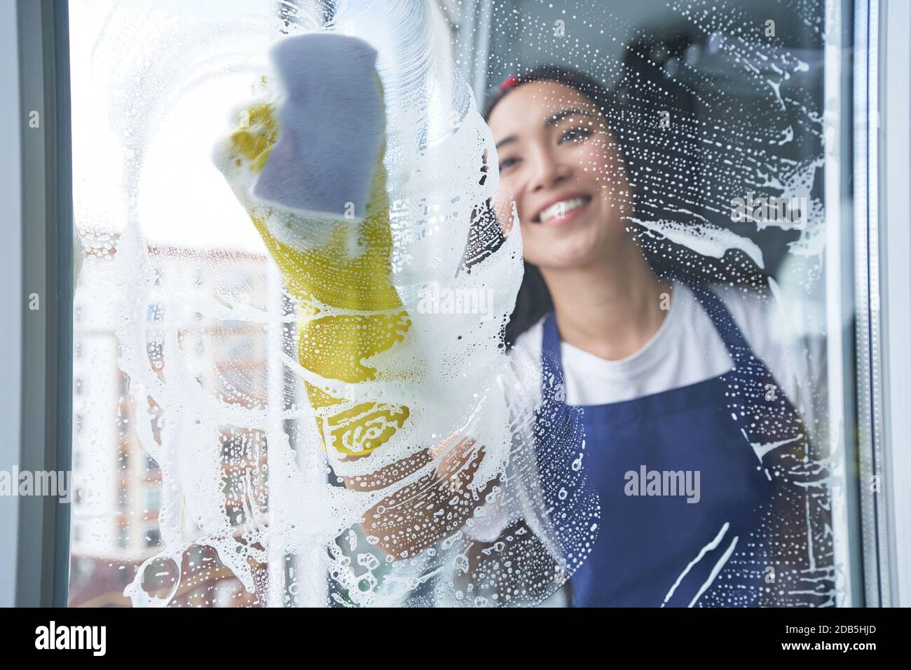 Fröhliche junge Frau lächelt beim Reinigen des Fensters, Glasoberfläche mit Schwamm. Hausarbeit und Housekeeping, Reinigungsdienstkonzept. Blick durch das Glas Stockfoto