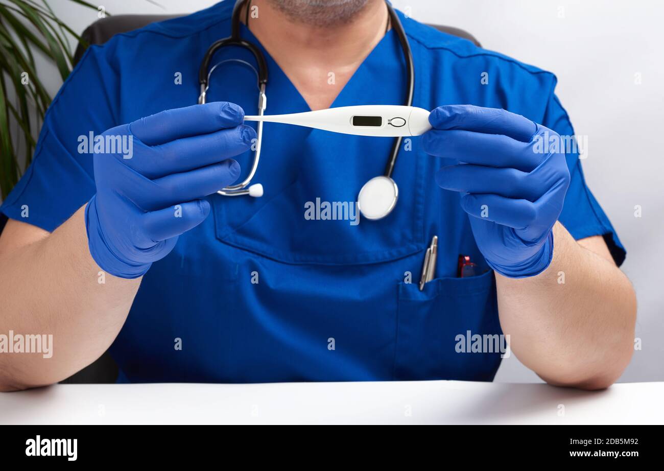 Doktor sitzt in blauer Uniform und Latexhandschuhe hält ein weißes elektronisches Thermometer, Arztpraxis Stockfoto