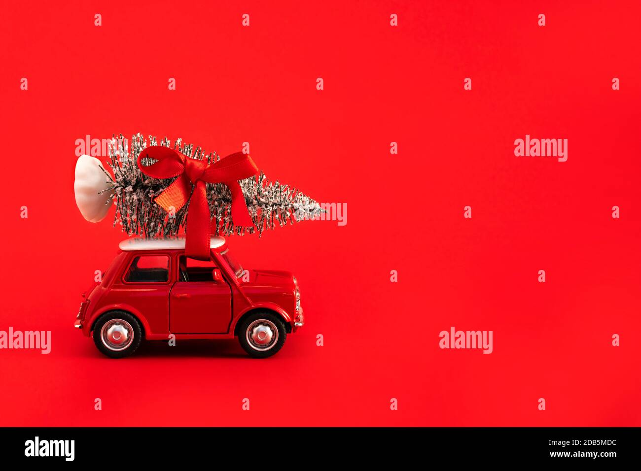 Rotes Spielzeug Auto trägt Weihnachtsbaum mit rotem Band auf dem Dach auf rotem Hintergrund. Platz für Text auf der rechten Seite. Stockfoto